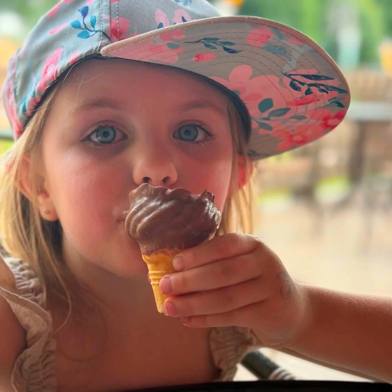 Petite fille avec une casquette rose et turquoise dégustant une crème glacée molle trempée dans le chocolat